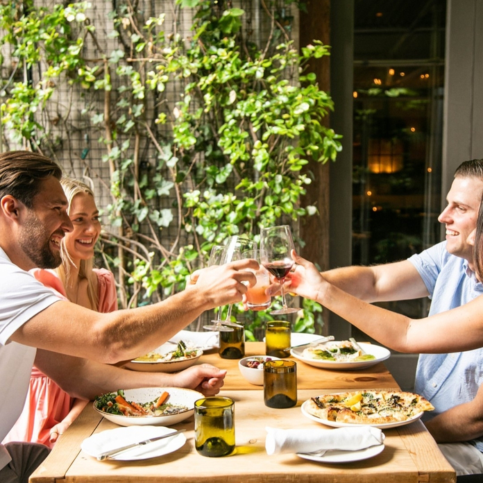 Three people celebrate with wine glasses at a wooden outdoor table with plates of food, surrounded by greenery.