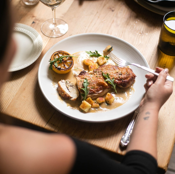 Dining scene with a person eating a plate of food featuring a meat dish, vegetables, and sauce on a wooden table with wine and beer glasses.