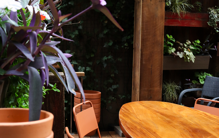Outdoor seating area with wooden table, terracotta chairs, and stacked pots. Plants and greenery add a natural ambiance.