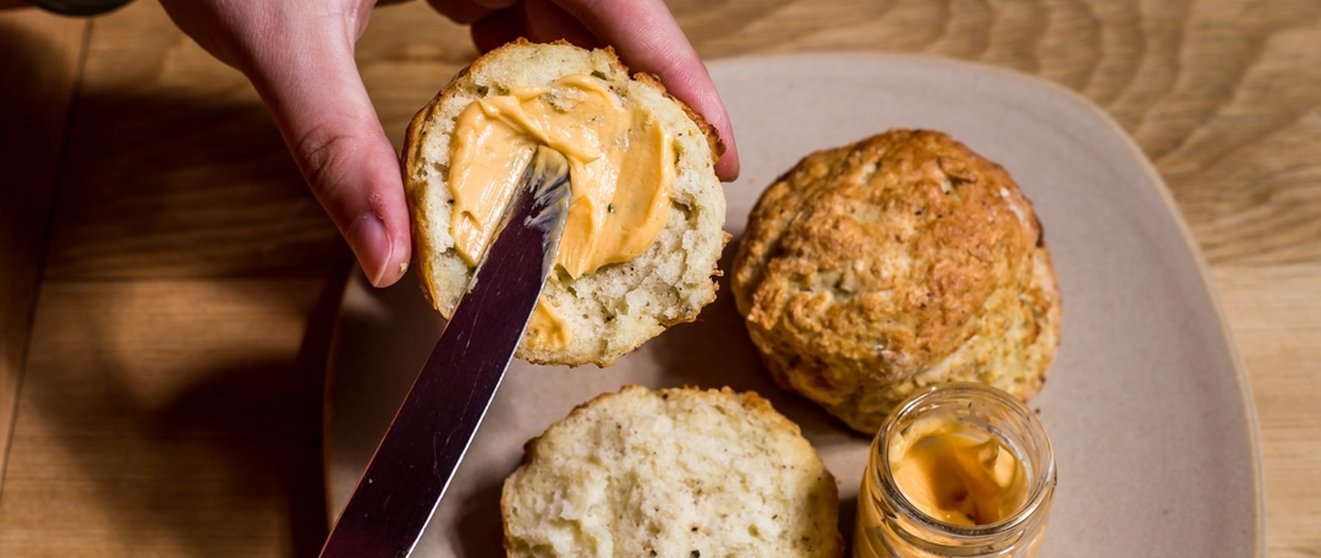 Hand spreading cheese on a biscuit on a beige plate with another biscuit and a jar of cheese.