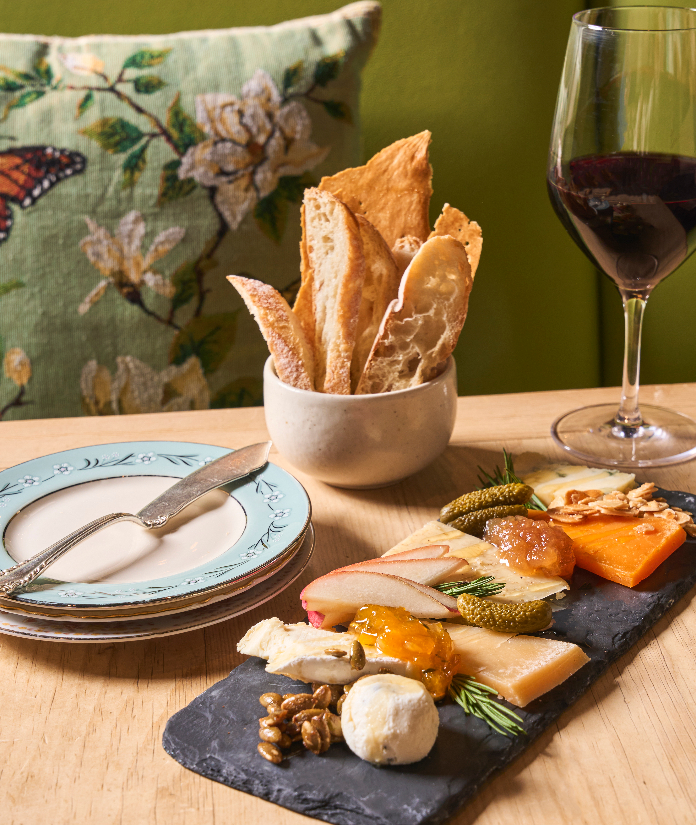 Charcuterie board with assorted cheeses, crackers in bowl, red wine glass, and decorative place setting on wooden table