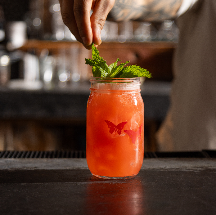A bartender garnishing a bright red cocktail in a mason jar with fresh mint, served on a dark bar counter with a butterfly design on the glass.