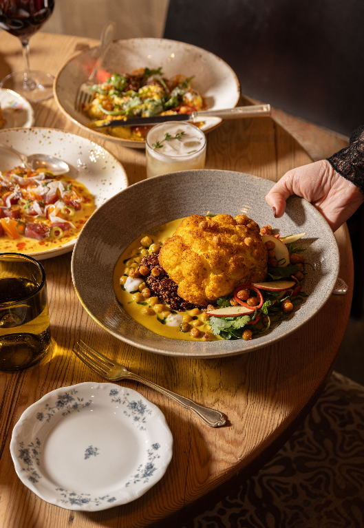 A server presenting a large bowl of golden cauliflower over grains and vegetables on a restaurant table, surrounded by other colorful dishes, drinks, and place settings.