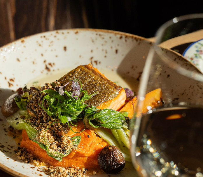 A close-up of a crispy-skinned salmon dish served over bright orange purée with greens, herbs, mushrooms, and crunchy garnish, with a glass of white wine in the foreground.