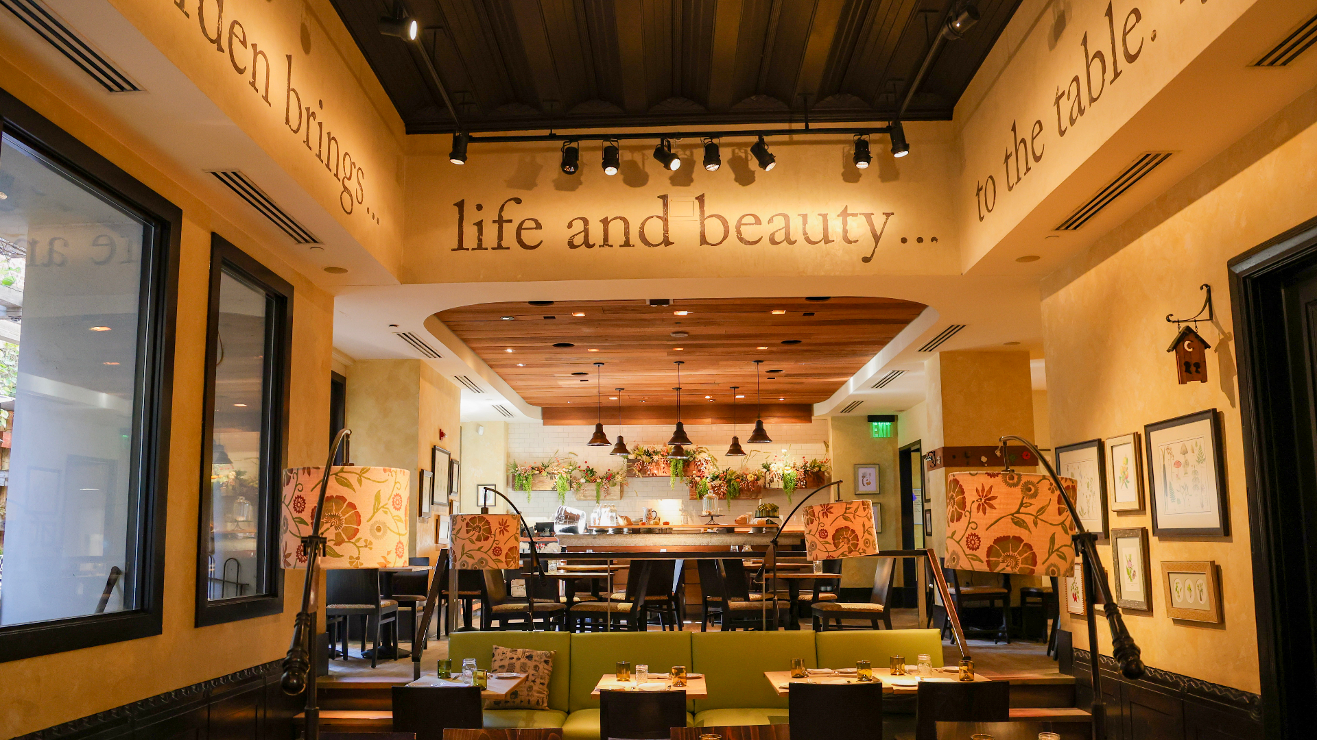 A warmly lit restaurant dining room with green booth seating, patterned chairs, framed art, and an open bar area in the back, with large wall text reading “life and beauty… to the table.”