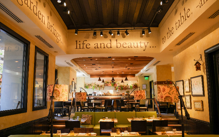 A warmly lit restaurant dining room with green booth seating, patterned chairs, framed art, and an open bar area in the back, with large wall text reading “life and beauty… to the table.”